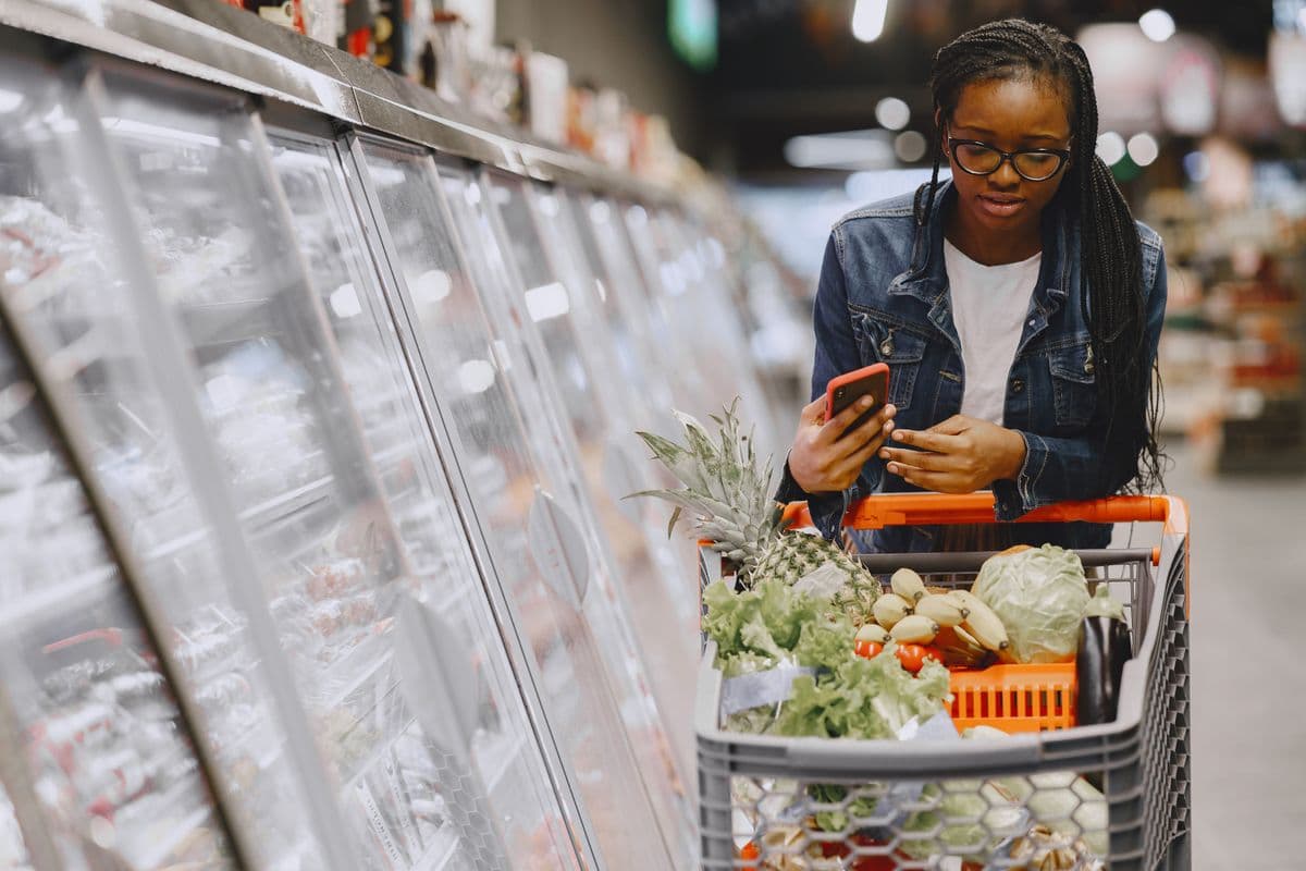 A person comparing prices in a grocery aisle, phone in hand, calculating a real weekly budget — not a styled market photo.